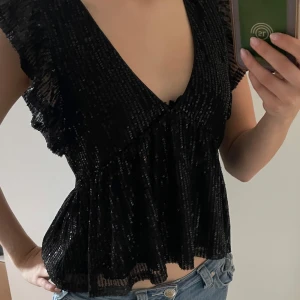 Svart glittrig topp från Nelly - Säljer en svart glittrig topp från Nelly med djup v-ringning och volangdetaljer vid axlarna. Toppen har en lätt utsvängd passform och är perfekt för att sticka ut. Materialet är syntet med paljetter som ger en snygg lyster.