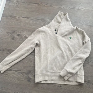 Ralph Lauren hoddie  - Säljer en beige hoodie från Polo Ralph Lauren med hög krage och knappar vid halsen. Tröjan har lång ärm och den klassiska gröna loggan broderad på bröstet. Perfekt för en avslappnad stil. Liten i storleken 