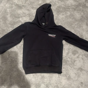 Svart hoodie från Balenciaga - Svart hoodie från Balenciaga med logga tryckt på bröstet och stor logga på ryggen. Klassisk känguruficka framtill och huva. Perfekt för en avslappnad och trendig stil.