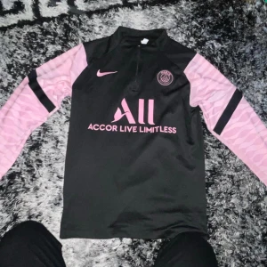 PSG långärmad fotbollströja svart/rosa - Säljer en långärmad Paris Saint-Germain fotbollströja från Nike. Tröjan är svart med rosa detaljer, rosa Nike-logga och PSG-märke på bröstet. Sponsorerna 'Accor Live Limitless' och 'Visit Rwanda' syns i rosa. Ärmarna är rosa med svart rand.