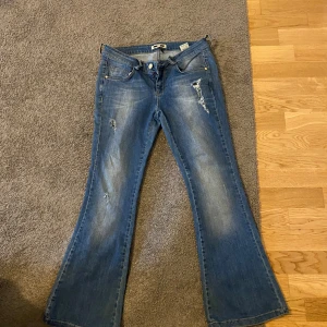 Låg midjade blå bootcut jeans med slitningar  - Säljer ett par blå bootcut jeans  med slitna detaljer och klassisk femficksdesign. Jeansen har normal passform och är tillverkade i denim med dragkedja och knapp framtill. Står 40 men passar som en 38