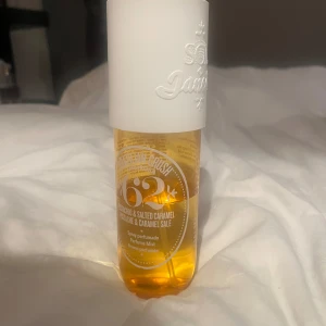 Brazilian Crush Cheirosa '62 Perfume Mist - En fräsch och söt body mist från Sol de Janeiro med doft av pistage och saltad karamell. Flaskan är genomskinlig med guldfärgad vätska och har ett stort vitt lock med präglad logga. Perfekt för dig som älskar gourmanddofter.