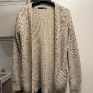 Beige stickad kofta från FB Sister - Mysig beige stickad kofta från FB Sister i storlek XS. Koftan har två fickor framtill och är öppen utan knäppning. Perfekt för lager-på-lager och passar till det mesta. Snyggt grovstickat material.