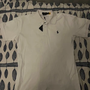 Vit piké från Polo Ralph Lauren - Klassisk vit pikétröja från Polo Ralph Lauren med broderad logga på bröstet. Tröjan har krage, knappar vid halsen och är kortärmad. Perfekt för en stilren och tidlös look.