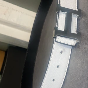 Svart och vit vändbart bälte med silverfärgat spänne - Stilrent vändbart bälte i svart och vitt med ett stort silverfärgat H-spänne. Bältet är gjort i skinn och har en längd på 110 cm, vilket motsvarar storlek L för herr. Pris kan diskuteras 