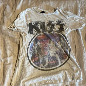 Vit KISS t-shirt från Gina Tricot - Säljer en vit t-shirt från Gina Tricot med stort KISS-tryck framtill. T-shirten har klassisk passform, rund hals och korta ärmar. Perfekt för dig som gillar bandtröjor och vill ha en cool och avslappnad stil.