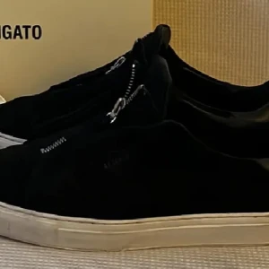 Svarta sneakers från Axel Arigato med dragkedja - Säljer ett par svarta sneakers från Axel Arigato med vit platt sula och dragkedja på ovansidan. Skorna har en stilren design och är tillverkade i mocka. Perfekta för dig som gillar minimalistisk stil.