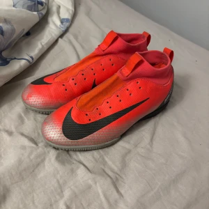 Nike Mercurial fotbollsskor i rött - Säljer ett par Nike Mercurial fotbollsskor i en stark röd färg med svart swoosh-logga. Skorna har en strumpliknande passform utan snörning och en låg profil, perfekt för snabba rörelser på planen. Snygg och modern design.