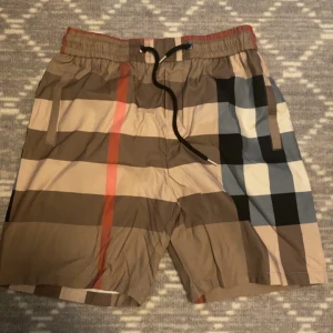Burberry shorts med klassiskt rutmönster - Snygga burberry shorts med klassiskt rutigt mönster i beige, svart, rött och blått. De har elastisk midja med svart snörning och sidofickor. Perfekta för sommaren och ger en trendig look.