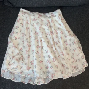 Vit blommig kjol - Charmig vit kjol med ett sött blommönster i rosa och grått från Nakd. Kjolen har en luftig design . Perfekt för varma dagar och somriga äventyr.