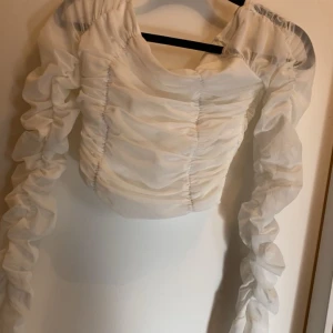 Vit blus med rynkade detaljer - Säljer en elegant vit blus med rynkade detaljer och långa ärmar. Blusen har en unik design med transparenta partier och en tight passform. Perfekt för en stilfull look. Den är från Nelly i storlek XS helt ny skick 
