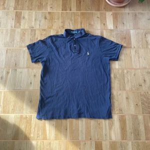 Blå pikétröja från Ralph Lauren - Säljer en klassisk blå pikétröja från Ralph Lauren med den ikoniska loggan broderad i gult på bröstet. Tröjan har en krage med två knappar och korta ärmar. Perfekt för en stilren look!