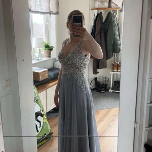 Grå långklänning med paljetter, balklänning - Elegant grå långklänning med djup v-ringning och öppen rygg. Överdelen är dekorerad med glittrande paljetter som ger en lyxig känsla. Perfekt för speciella tillfällen.