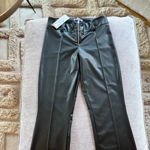 Svarta skinnbyxor med dragkedja - Snygga svarta skinnbyxor med låg midja och dragkedja framtill. Byxorna har en stilren design med raka ben och markerade sömmar längs benen. Perfekta för en trendig look.