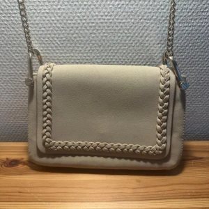 Beige axelväska med flätad detalj - Snygg beige axelväska med flätad kantdetalj och kedjeaxelrem. Perfekt för att ge en stilren touch till din outfit. Väskan har en elegant design och är tillverkad i ett mjukt material.
