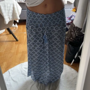 Blå mönstrad långkjol - Säljer en blå långkjol med ett snyggt mönster i vitt och mörkblått. Kjolen har en elastisk midja och dekorativa tofsar framtill. Använt bara ett fåtal gånger, säljer den då den är för kort för mig😊