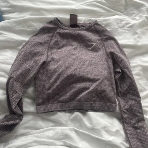 Lila-Gråmelerad långärmad tröja från Gymshark - Säljer en gråmelerad långärmad tröja från Gymshark. Tröjan har en tight passform och är tillverkad i ett syntetmaterial som andas. Perfekt för träning eller en aktiv vardag. Den har en diskret logga på bröstet och en rund halsringning.