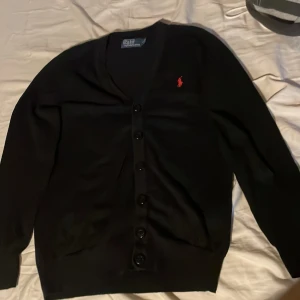 Svart kofta från Polo Ralph Lauren - Snygg svart kofta från Polo Ralph Lauren med knappar framtill och det ikoniska röda logotypen på bröstet. Perfekt för en stilren look.