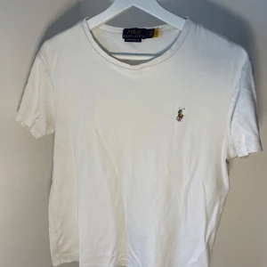 Vit t-shirt från Polo Ralph Lauren - Säljer en klassisk vit t-shirt från Polo Ralph Lauren med broderad logga på bröstet. T-shirten har en slim fit passform och är tillverkad i mjuk bomull. Perfekt för en stilren och avslappnad look. Tröjan har ett litet litet hål vid slutet av framsidan (se bild 3). Dessutom små spår av missfärgning (bild 4). Annars i fint skick.