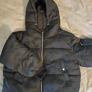 Camouflage pufferjacka från Zara - Säljer en cool camouflage pufferjacka från Zara Basic. Jackan har en dragkedja framtill och praktiska fickor med knappar. Den är perfekt för kyliga dagar med sin vadderade design och har en huva för extra skydd. Muddar vid ärmsluten håller kylan ute. Perfekt för dig som vill ha en stilren look med en edge. Lite nopprig på muddar 