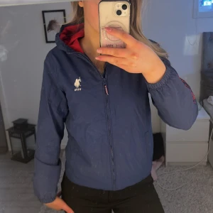 Blå vindjacka från U.S. Polo Assn. - Snygg blå jacka från U.S. Polo Assn. med dragkedja och röd insida💙sparsamt använd väldigt fint skick! Spegeln är lortig ej jackan!