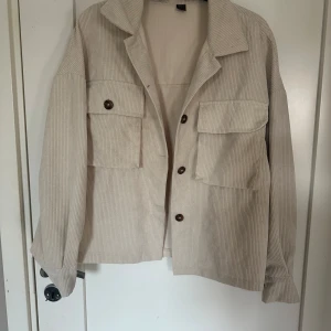 Beige overshirt med knappar - Säljer en stilren beige overshirt med knappar framtill och två stora fickor på bröstet. Jackan har en avslappnad passform och är perfekt för lager-på-lager-stil. Den har långa ärmar och en klassisk krage.