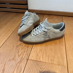 Grå sneakers med från Scalpers  - Snygga grå sneakers i mocka med ett coolt dödskallemotiv på sidan. Skorna har en brun sula och snörning framtill. Perfekta för en avslappnad stil. Knappt använda i toppen skick