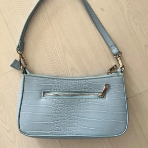 Ljusblå axelväska  - Snygg ljusblå axelväska med krokodilmönster och dragkedja framtill. Perfekt för att ge din outfit en trendig touch. Väskan har en justerbar axelrem och en praktisk ficka med dragkedja för enkel förvaring.