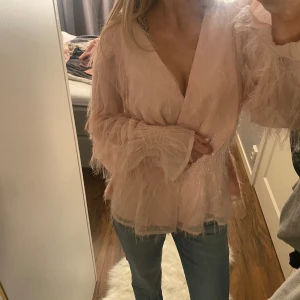 Rosa blus med fjäderdetaljer - Säljer en unik rosa blus med fjäderdetaljer och v-ringning. Blusen har långa ärmar och ett omlottband i midjan som ger en elegant look. Perfekt för att sticka ut med en touch av glamour.