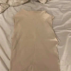 Beige ärmlös topp från Zara - Säljer en stilren beige ärmlös topp från Zara. Perfekt för lager-på-lager eller att bära som den är. Toppen har en ribbad textur som ger en extra touch av stil. Passar till många olika outfits!