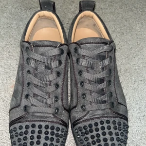 Grå sneakers med nitar från Christian Louboutin - Snygga grå sneakers från Christian Louboutin med svarta nitar på tån och den ikoniska röda sulan. Skorna har snörning och är tillverkade i ett stilrent material som ger en lyxig känsla. Perfekta för den som vill sticka ut med stil.  PRIS KAN DISKUTERAS!