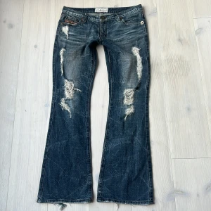 Blå bootcut jeans  - Snygga blå bootcut jeans med slitna detaljer och en låg midja. De har en cool vintage-look med slitningar på framsidan och baksidan. Storlek 28. Midjemått: 41cm tvärs över, innerbenslängden: 83cm. Små slitningar längst ner vid hälen, se bild.