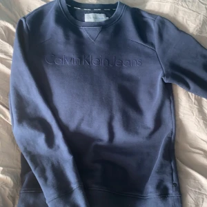 Blå sweatshirt från Calvin Klein Jeans - Säljer en stilren blå sweatshirt från Calvin Klein Jeans. Tröjan har en diskret logga på bröstet och ribbade muddar vid ärmslut och nederkant. Perfekt för en avslappnad look. Pris kan diskuteras vid snabb affär