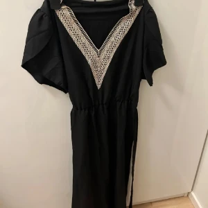 Svart klänning med spetsdetaljer - Elegant svart klänning med v-ringad spetsdetalj i beige. Klänningen har korta ärmar och en avslappnad passform. Perfekt för en stilren look.