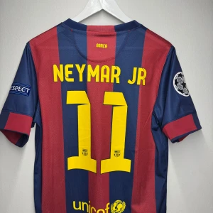 FC Barcelona 2014/2015 Stadium (hemmaställ) - Säljer en FC Barcelona fotbollströja med Neymar Jr på ryggen. Den har Champions League-märken på ärmarna (inte använd utan bara upphängd på en galje i mitt rum) Tröjan är väldigt unik och exklusiv ifrån barcelonas Treble år. (Ingen aning om hur äkta den är men kvalitèn är sjukt obeskrivlig som ni ser och samma med all dess material)
