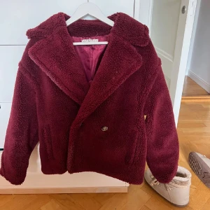 Vinröd teddyjacka från Vila - Mysig vinröd teddyjacka från Vila med knappar framtill. Jackan har en klassisk krage och är perfekt för kyliga dagar. Den är fodrad med ett matchande tyg på insidan för extra värme.