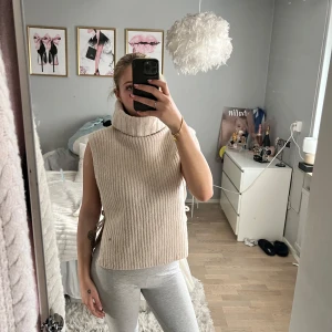 Beige ribbad ärmlös polotröja -  beige ribbad polotröja utan ärmar. Perfekt för lager-på-lager eller som den är. Tröjan har en hög krage och en bekväm passform. Perfekt för både höst och vår.