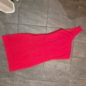Röd one-shoulder klänning - Snygg röd one-shoulder klänning med dragkedja i sidan. Perfekt för en kväll ute eller en speciell tillställning. Klänningen har en figurnära passform som framhäver silhuetten. Endast använd 1 gång.