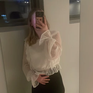 Vit blus med volangdetaljer - Säljer en elegant vit blus med genomskinliga ärmar och volangdetaljer. Blusen har en rund hals och knäppning vid ärmsluten. Perfekt för en stilren look. Bandet för att knäppa på baksidan är sönder och går lätt att sy ihop men jag har använt den ändå och det har funkat perfekt. Går att buda 