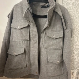 Grå jacka från Gina Tricot - Snygg grå jacka från Gina Tricot med dragkedja och knappar. Jackan har flera praktiska fickor framtill och en hög krage. Den är i väldigt bra skick och köptes för 599.