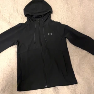 Svart vindjacka från Under Armour - Säljer en stilren svart vindjacka från Under Armour. Jackan har en dragkedja framtill och en justerbar huva. Perfekt för blåsiga dagar och sportiga aktiviteter. Den är helt oanvänd🖤