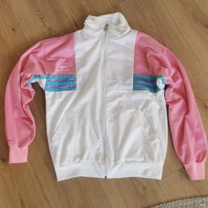 Vit och rosa vindjacka från Bagheera - Säljer en retro vit och rosa vindjacka från Bagheera med turkosa ränder på ärmarna. Jackan har dragkedja och långa ärmar. Perfekt för en sportig look! Jättefint skick förutom en liten reva på ärmen. Se bild! 
