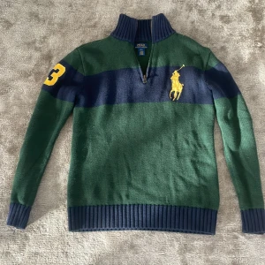 Grön och marinblå tröja från Ralph Lauren - Säljer en snygg grön och marinblå tröja från Ralph Lauren med dragkedja och ribbade muddar. Tröjan har ett stort gult logotryck på bröstet och en gul siffra på ärmen. Perfekt för en stilren look!