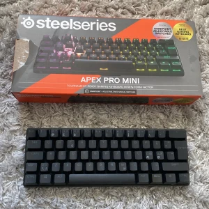 SteelSeries Apex Pro Mini Gamingtangentbord - Säljer ett SteelSeries Apex Pro Mini gamingtangentbord i 60% formfaktor. Utrustat med OmniPoint justerbara mekaniska switchar och RGB-belysning. Perfekt för turneringsspelare som vill ha snabb respons och kompakt design. Inkluderar en orange keycap-puller för enkel anpassning. Höj och sänkbart och trådbundet spel tangentbord. Endast en liten skada på boxen men ingen skada på tangentbordet.