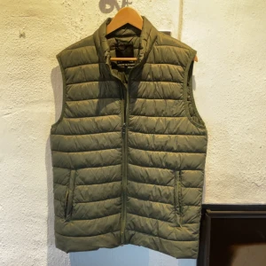 Olivgrön quiltad dunväst - Snygg olivgrön quiltad dunväst med dragkedja framtill. Perfekt för lager-på-lager under kyliga dagar. Västen har en stilren design med två fickor på sidorna. Använd ca 10 gånger.                                      Vid frågor och funderingar hör gärna av er!                                                                            Mvh Doré.reselll