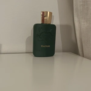 Haltane Parfym från Parfums de Marly - Elegant parfymflaska i mörkgrönt med guldfärgat lock. Flaskan har en stilren design med två hästar i relief och texten 'Haltane'. Perfekt för den som söker en sofistikerad doftupplevelse. Ungefär 90-100ml kvar.