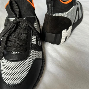 Svarta och vita sneakers med orange detaljer  - Snygga sneakers i svart och vitt med orange detaljer. Skorna har en stickad överdel med snörning och en robust sula. Perfekta för en stilren look med en touch av färg.
