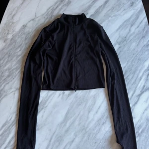 Svart ribbad tröja med dragkedja - Snygg svart ribbad tröja med dragkedja framtill som man kan öppna både uppifrån och nerifrån. Tröjan har en croppad design och långa ärmar, perfekt för en stilren look. 