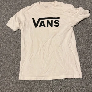 Vit t-shirt från Vans 10-12  - Säljer en klassisk vit t-shirt från Vans med deras ikoniska logga i svart på bröstet. T-shirten är tillverkad i 100% bomull och har en normal passform. Perfekt för en avslappnad stil. Size=10-12