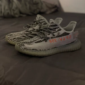Adidas Yeezy Boost 350 V2 Zebra - Säljer ett par Adidas Yeezy Boost 350 V2 i den populära Zebra-färgen. Skorna har en unik randig design i svart och vit med röd text på sidan. De är kända för sin bekväma passform och stilrena look.